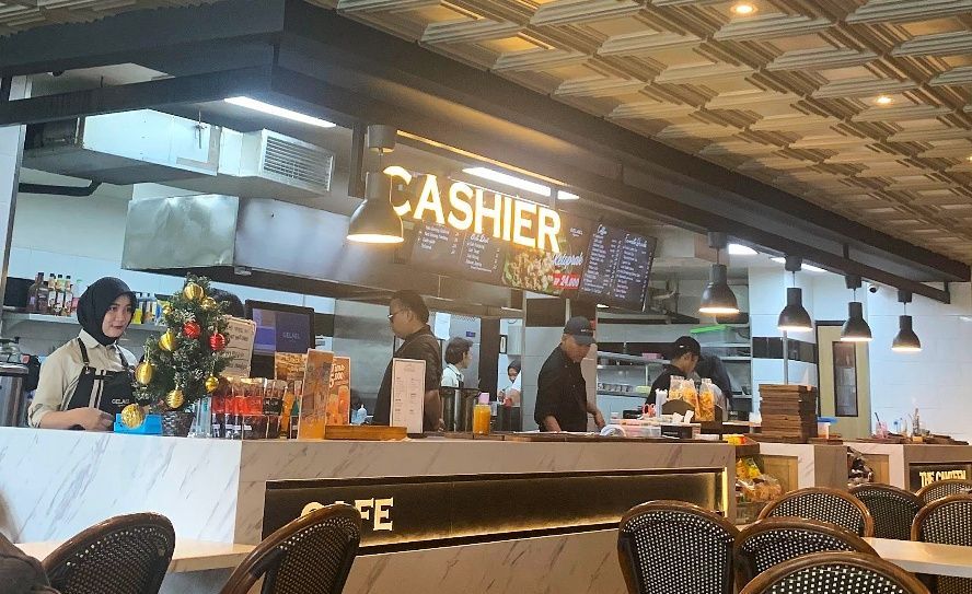 Petugas kasir wanita berhijab melayani pelanggan di area meja kasir Gelael dengan latar belakang staf dapur sedang menyiapkan makanan.