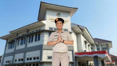 Seorang pegawai pria berseragam khaki dan berpeci hitam melakukan salam dengan tangan ditangkupkan di depan gedung Kantor Pertanahan Kota Semarang di bawah langit biru yang cerah.
