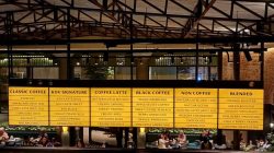 Papan menu besar berwarna kuning di KOV Heritage Semarang yang menampilkan daftar kopi signature, latte, dan non-coffee dengan latar belakang interior industrial cagar budaya.