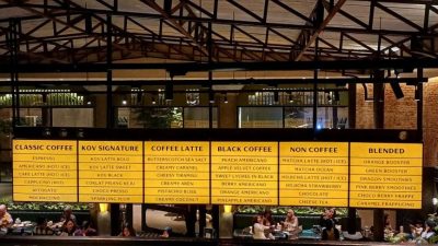 Papan menu besar berwarna kuning di KOV Heritage Semarang yang menampilkan daftar kopi signature, latte, dan non-coffee dengan latar belakang interior industrial cagar budaya.