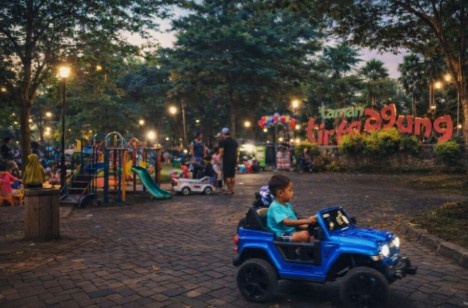 Anak bermain mobil mainan di Taman Tirto Agung Banyumanik saat sore hari dengan latar playground, pepohonan rindang, dan suasana taman kota Semarang.