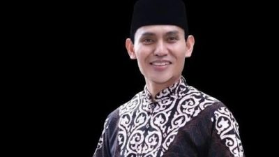 Potret Ketua Umum DPP BKPRMI KH. Nanang Mubarok mengenakan baju koko gelap bermotif putih dan peci hitam dengan latar belakang hitam.