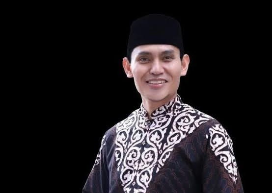 Potret Ketua Umum DPP BKPRMI KH. Nanang Mubarok mengenakan baju koko gelap bermotif putih dan peci hitam dengan latar belakang hitam.